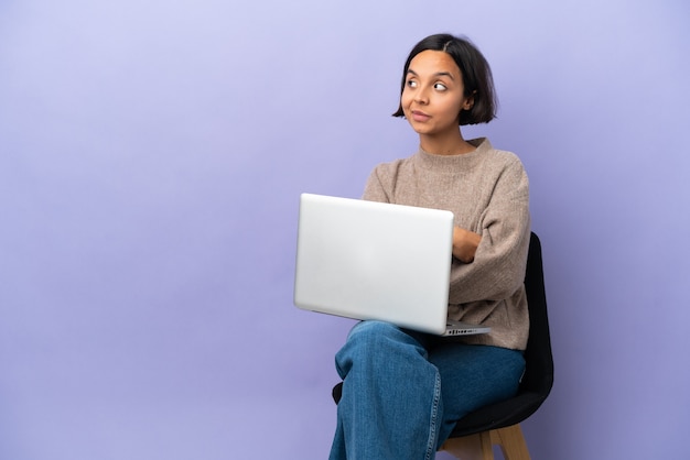 Jonge gemengd ras vrouw zittend op een stoel met laptop geïsoleerd op paarse achtergrond met gekruiste armen en happy