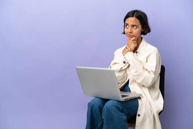 Jonge gemengd ras vrouw zittend op een stoel met laptop geïsoleerd op paarse achtergrond met twijfels en met verwarde gezichtsuitdrukking