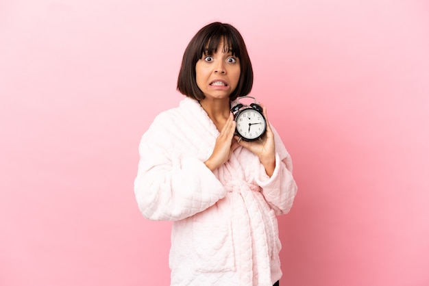 Jonge gemengd ras zwangere vrouw geïsoleerd op roze achtergrond in pyjama en klok met verbaasde uitdrukking te houden