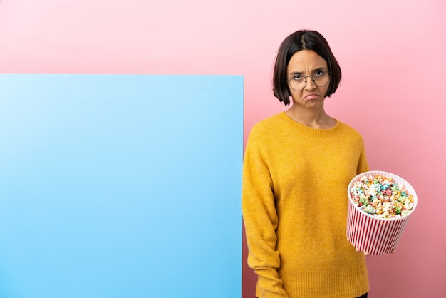 Jonge gemengde race vrouw met popcorn met een grote banner over geïsoleerde achtergrond met droevige uitdrukking