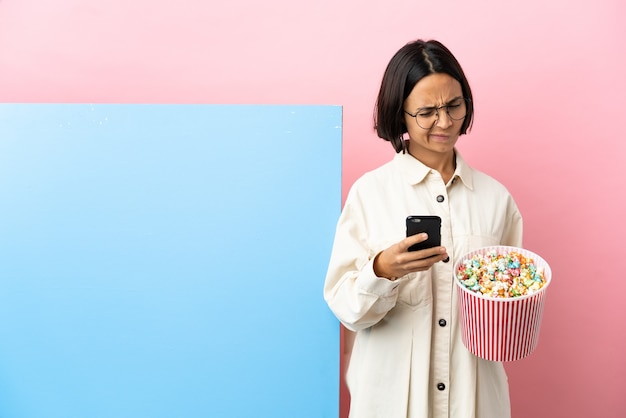 Jonge gemengde race vrouw met popcorns met een grote banner over geïsoleerde achtergrond denken en verzenden van een bericht