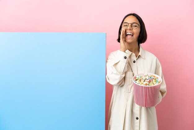 Jonge gemengde race vrouw met popcorns met een grote banner over geïsoleerde achtergrond schreeuwen en iets aankondigen