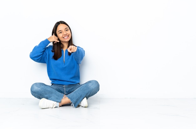 Jonge gemengde race vrouw zittend op de vloer geïsoleerd op wit telefoon gebaar maken en naar voren wijzen