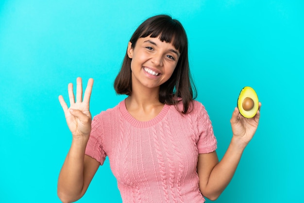 Jonge gemengde rasvrouw die een avocado houdt die op blauwe achtergrond gelukkig wordt geïsoleerd en vier met vingers telt