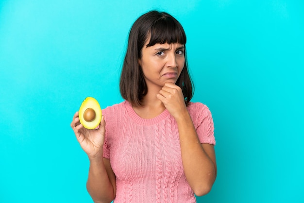 Jonge gemengde rasvrouw die een avocado houdt die op blauwe achtergrond wordt geïsoleerd die twijfels heeft