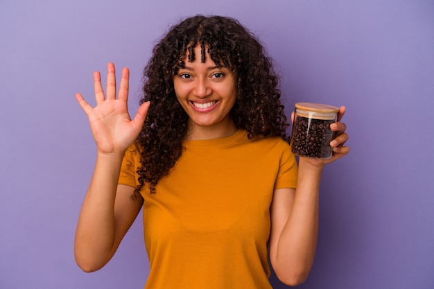 Jonge gemengde rasvrouw die een fles van koffiebonen houden die op purpere muur wordt geïsoleerd vrolijk glimlachend tonend nummer vijf met vingers.