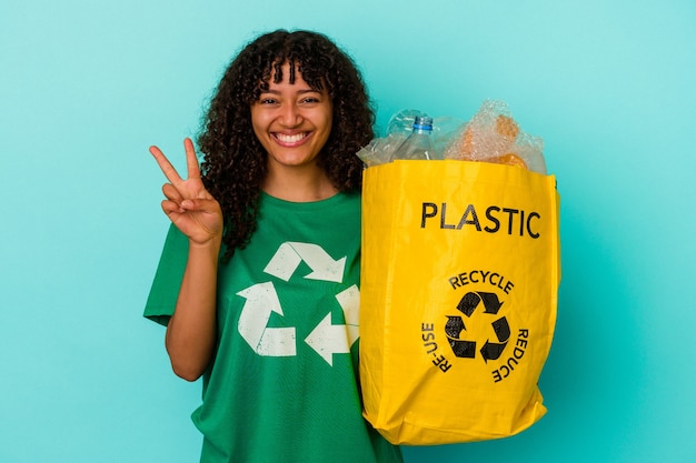 Jonge gemengde rasvrouw die een gerecycleerde plastic zak houdt die op blauwe achtergrond wordt geïsoleerd die nummer twee met vingers toont.