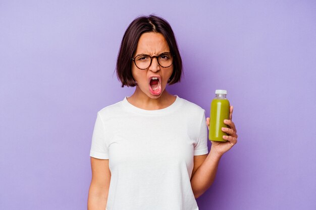 Jonge gemengde rasvrouw die een gezonde smoothie houden die op purpere achtergrond wordt geïsoleerd die zeer boos en agressief schreeuwen.