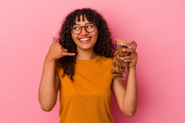 Jonge gemengde rasvrouw die een koekjeskruik houden die op roze muur wordt geïsoleerd die een mobiel telefoongesprekgebaar met vingers toont.
