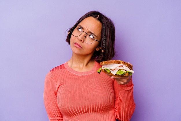 Jonge gemengde rasvrouw die een sandwich houden die op purpere achtergrond wordt geïsoleerd die dromen van het bereiken van doelstellingen en doeleinden