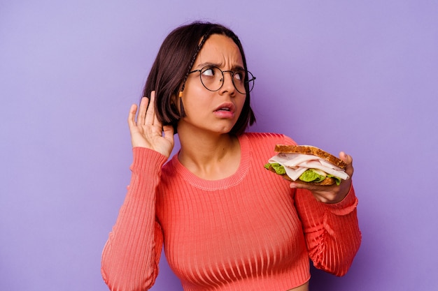 Jonge gemengde rasvrouw die een sandwich houdt die op purpere achtergrond wordt geïsoleerd die probeert een roddel te luisteren.