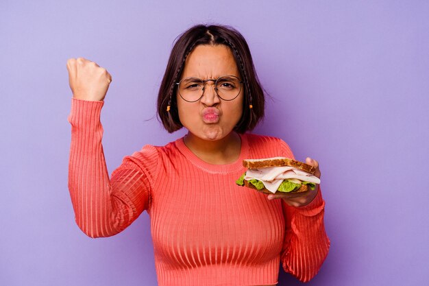 Jonge gemengde rasvrouw die een sandwich houdt die op purpere achtergrond wordt geïsoleerd die vuist toont aan camera, agressieve gezichtsuitdrukking.