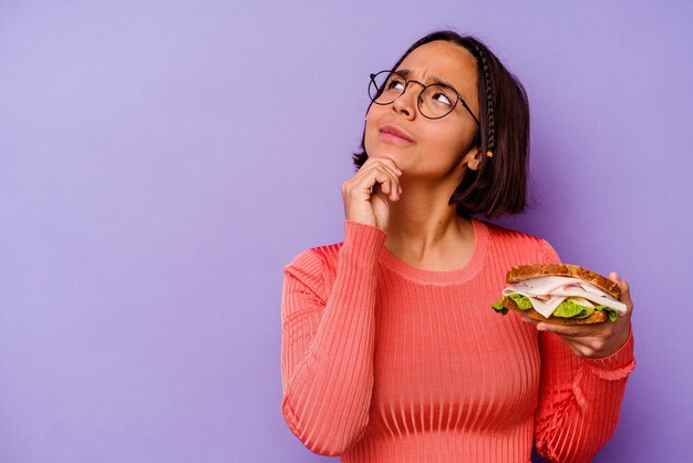 Jonge gemengde rasvrouw die een sandwich houdt die op purpere achtergrond wordt geïsoleerd die zijwaarts met twijfelachtige en sceptische uitdrukking kijkt.