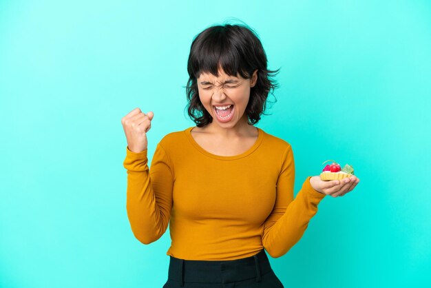 Jonge gemengde rasvrouw die een taartje houdt dat op blauwe achtergrond wordt geïsoleerd en een overwinning viert