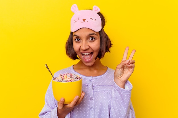 Jonge gemengde rasvrouw die granen eet die een pijama dragen die op gele muur wordt geïsoleerd vrolijk en zorgeloos met een vredessymbool met vingers.