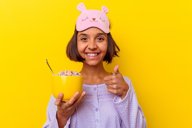 Jonge gemengde rasvrouw die granen eten die een pijama dragen die op gele achtergrond wordt geïsoleerd die en duim glimlacht opheft