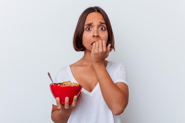 Jonge gemengde rasvrouw die granen eten die op witte muur worden geïsoleerd die vingernagels bijten, zenuwachtig en zeer angstig.