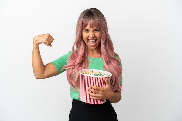 Jonge gemengde rasvrouw die met roze haar popcorn eet die op witte achtergrond wordt geïsoleerd en sterk gebaar doet