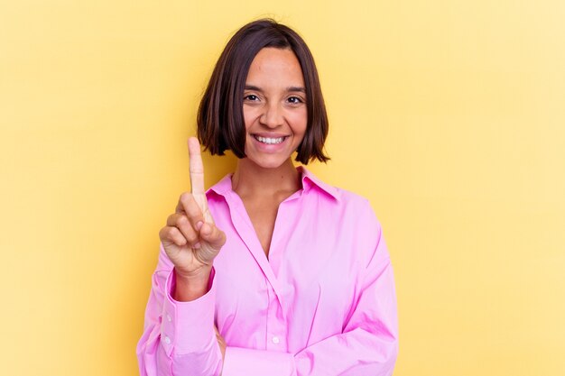 Jonge gemengde rasvrouw die op gele achtergrond wordt geïsoleerd die nummer één met vinger toont.