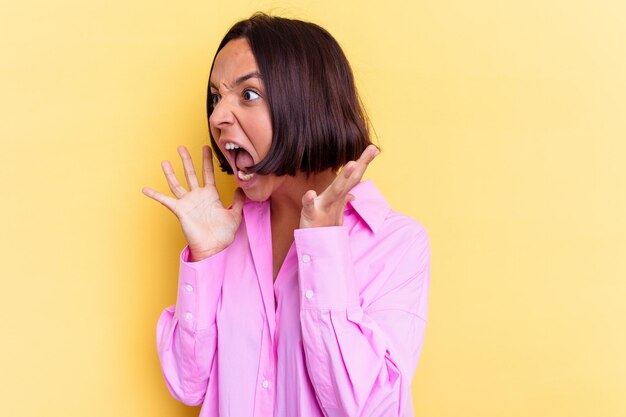 Jonge gemengde rasvrouw die op gele muur wordt geïsoleerd schreeuwt luid, houdt ogen open en handen gespannen.