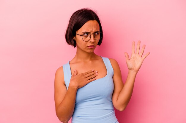 Jonge gemengde rasvrouw die op roze wordt geïsoleerd die een eed aflegt, die hand op borst legt.