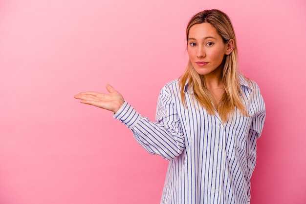 Jonge gemengde rasvrouw die op roze wordt geïsoleerd die een exemplaarruimte op een palm toont en een andere hand op taille houdt.