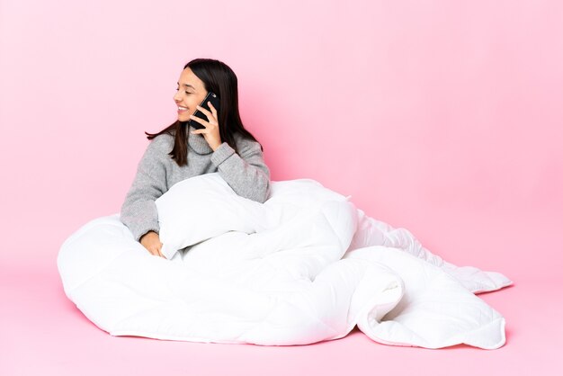 Jonge gemengde rasvrouw die pijama draagt die op de vloer zit die een gesprek met de mobiele telefoon met iemand houdt