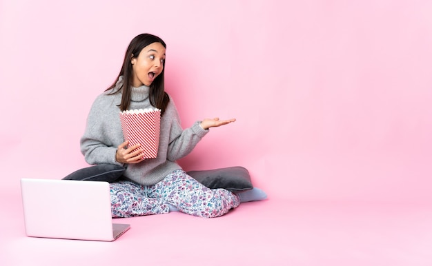 Jonge gemengde rasvrouw die popcorn eet tijdens het kijken naar een film op de laptop die een idee presenteert terwijl ze glimlachend naar kijkt