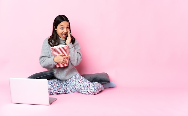 Jonge gemengde rasvrouw die popcorn eet tijdens het kijken naar een film op de laptop die naar de zijkant wijst om een product te presenteren en iets fluistert