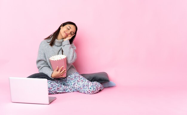 Jonge gemengde rasvrouw die popcorn eet tijdens het kijken naar een film op de laptop die slaapgebaar maakt in heerlijke uitdrukking