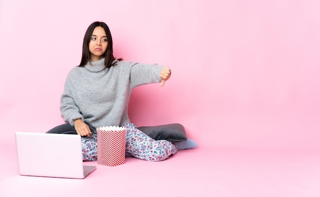 Jonge gemengde rasvrouw die popcorn eet tijdens het kijken naar een film op de laptop met duim omlaag met negatieve uitdrukking