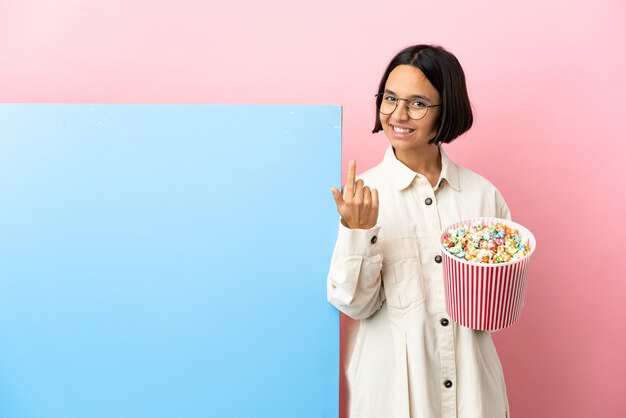 Jonge gemengde rasvrouw die popcorns met een grote banner over geïsoleerde achtergrond houdt die komend gebaar doet