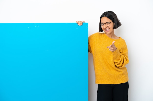 Jonge gemengde rasvrouw met een groot blauw aanplakbiljet dat op witte achtergrond wordt geïsoleerd die naar voren wijst en glimlacht