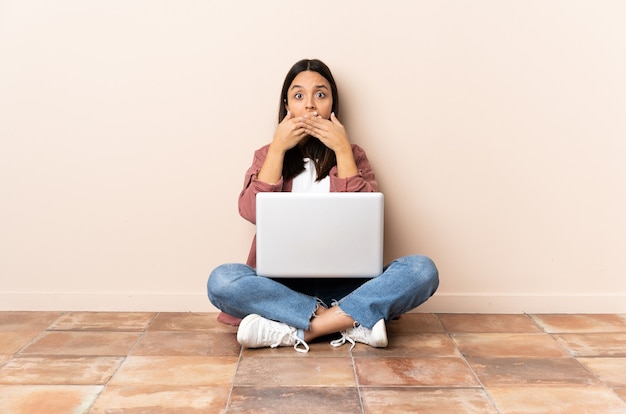 Jonge gemengde rasvrouw met laptopzitting op de vloer die mond met handen bedekken