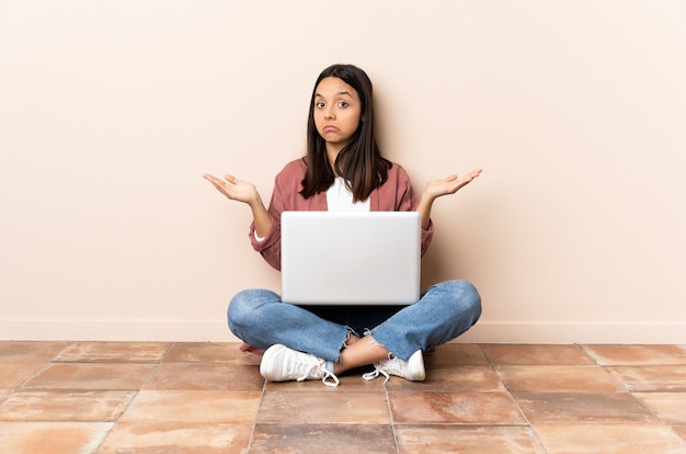 Jonge gemengde rasvrouw met laptopzitting op de vloer die twijfelsgebaar maken