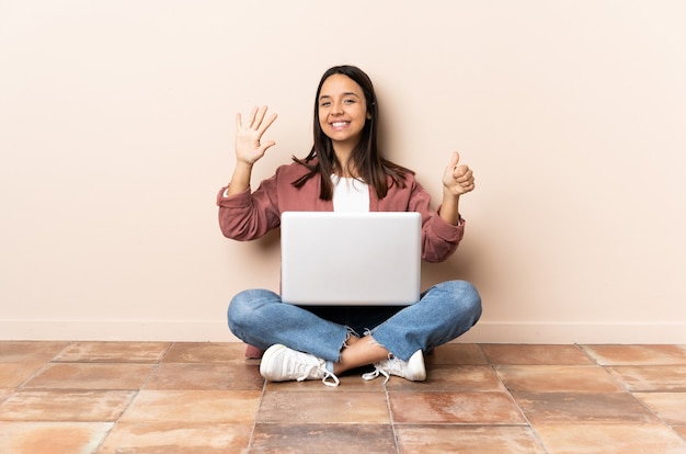 Jonge gemengde rasvrouw met laptopzitting op de vloer die zes met vingers telt