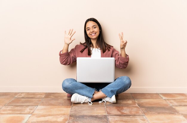 Jonge gemengde rasvrouw met laptopzitting op de vloer die zeven met vingers telt