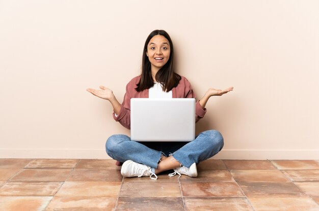 Jonge gemengde rasvrouw met laptopzitting op de vloer met geschokte gezichtsuitdrukking