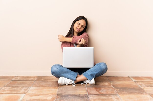 Jonge gemengde rasvrouw met laptopzitting op de vloer met pijn in elleboog