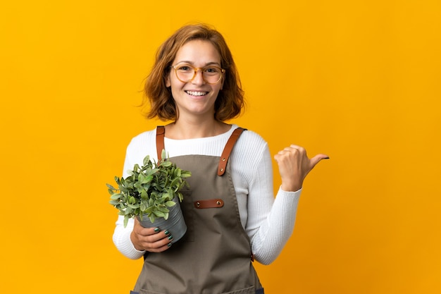 Jonge georgische vrouw die een plant houdt die op gele achtergrond wordt geïsoleerd die naar de kant wijst om een product te presenteren