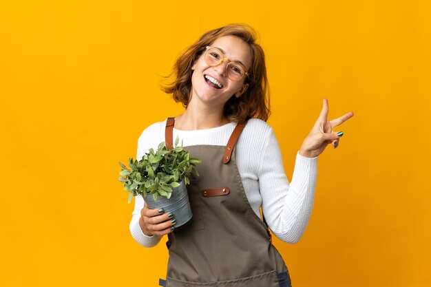 Jonge Georgische vrouw die een plant houdt die op gele muur wordt geïsoleerd die en overwinningsteken glimlacht toont