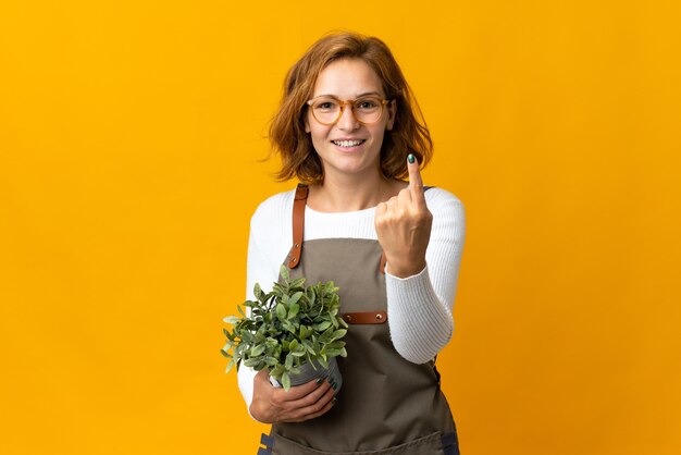 Jonge Georgische vrouw die een plant houdt die op gele muur wordt geïsoleerd die komend gebaar doet