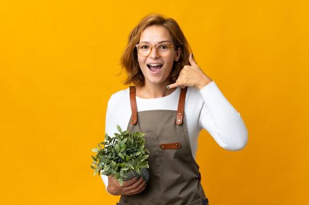 Jonge Georgische vrouw die een plant houdt die op gele muur wordt geïsoleerd die telefoongebaar maakt. Bel me terug teken