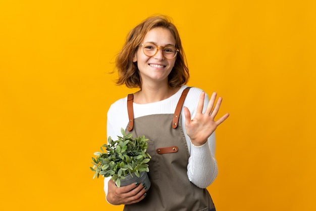 Jonge Georgische vrouw die een plant houdt die op gele muur wordt geïsoleerd die vijf met vingers telt