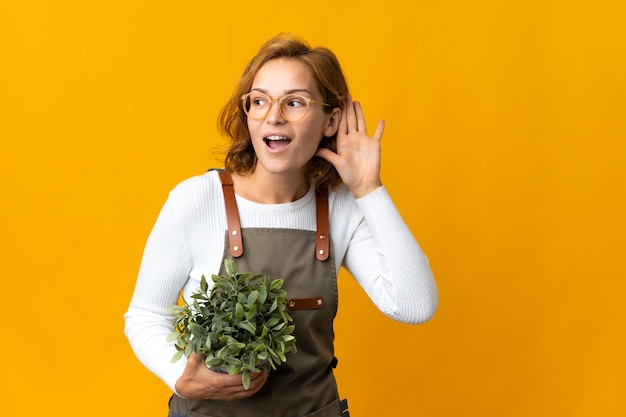 Foto jonge georgische vrouw die een plant vasthoudt die op een gele achtergrond is geïsoleerd en naar iets luistert door de hand op het oor te leggen