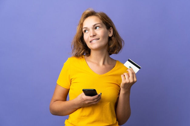Jonge Georgische vrouw geïsoleerd op paarse achtergrond kopen met de mobiele telefoon met een creditcard terwijl u denkt