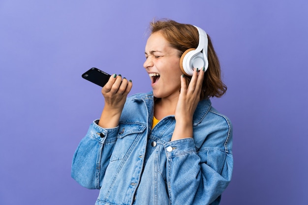 Jonge Georgische vrouw geïsoleerd op paarse achtergrond muziek luisteren met een mobiele telefoon en zingen