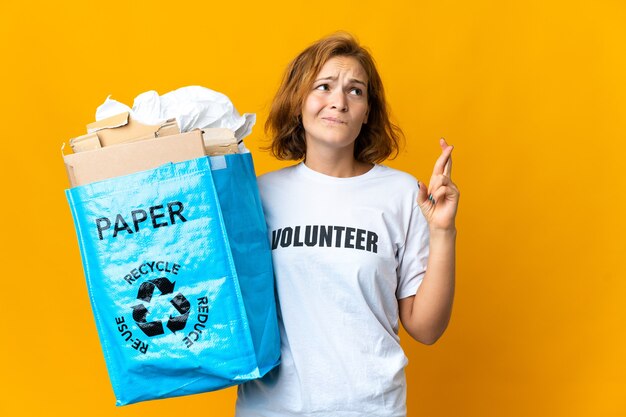 Jonge Georgische vrouw met een recyclingzak vol papier om te recyclen met gekruiste vingers en het beste wensen