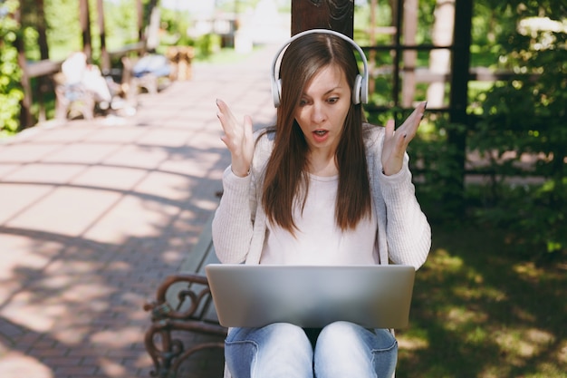 Jonge geschokte mooie zakenvrouw in lichte vrijetijdskleding. Vrouw die werkt op moderne laptop pc-computer, luister muziek in koptelefoon op hoofd in straat buitenshuis. Mobiel kantoor. Freelance bedrijfsconcept