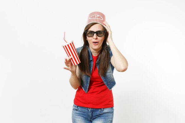 Jonge geschokte vrouw in 3D-bril met emmer voor popcorn op hoofd kijken naar film, met plastic beker frisdrank of cola, klampt zich vast aan hoofd geïsoleerd op een witte achtergrond. Emoties in bioscoopconcept.